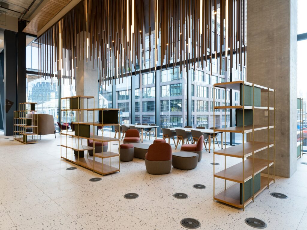 A modern open-concept workspace with large glass windows allowing natural light to fill the area. Sustainable wooden shelves and minimalist furniture create a flexible seating arrangement. Overhead, hanging wooden beams and lights add visual interest, emphasizing eco-conscious design.