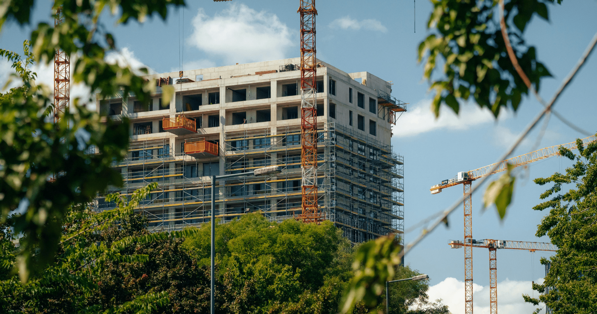 Mid-rise concrete building under construction with scaffolding and cranes, surrounded by trees.