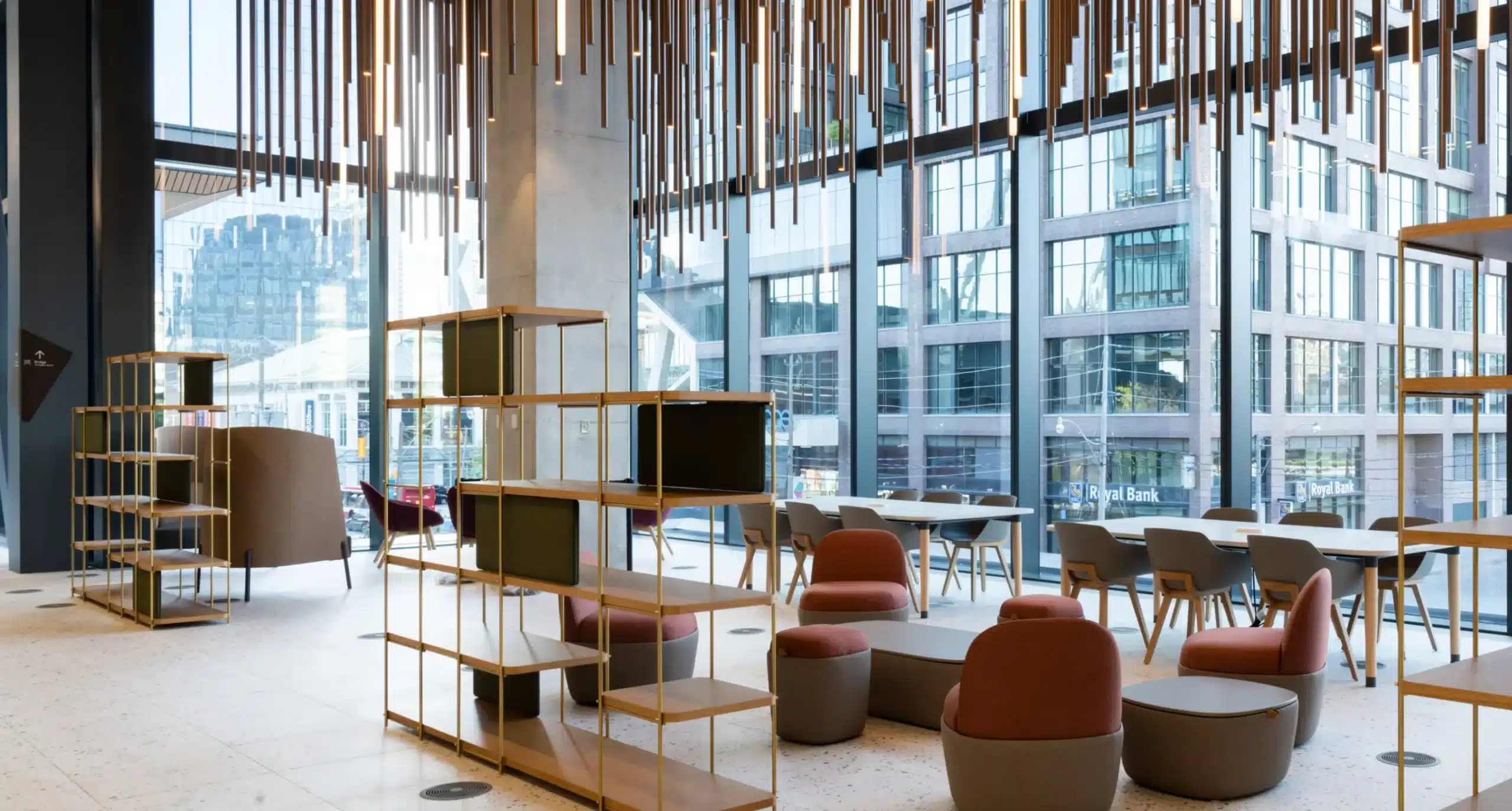 Modern office lounge with tables, chairs, shelving, and large floor-to-ceiling windows.